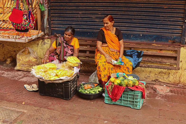 two women sit outside on their knees near some fruit