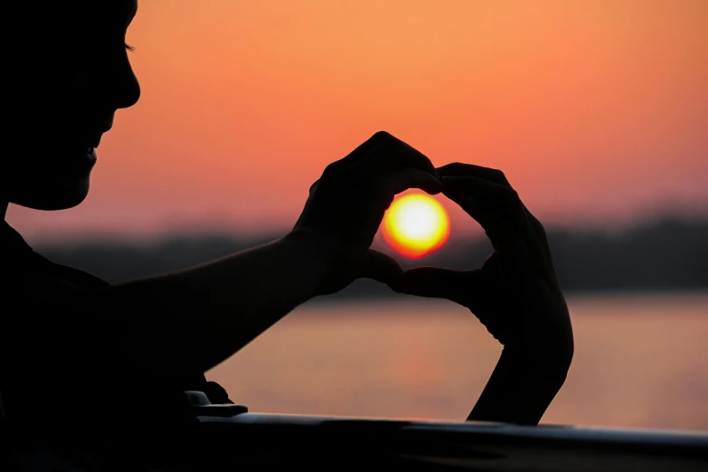 a person making a heart sign with their hand while sitting in front of a sunset