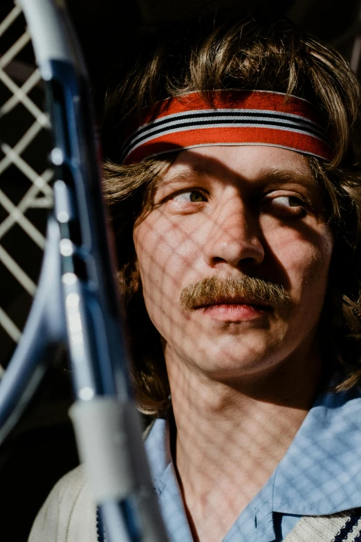 a man with a moustache, mustache and head band holds tennis racket in one hand and looks at the camera