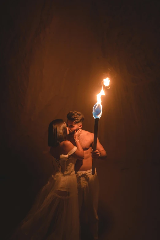 a man in a white dress kisses a girl's forehead, holding a stick lit up in flames