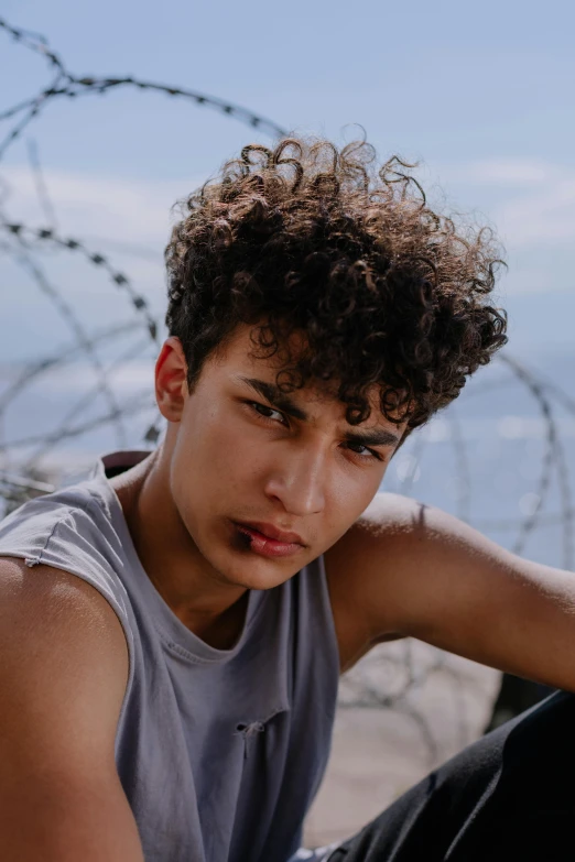 a young man with curly hair sitting on the ground by a fence