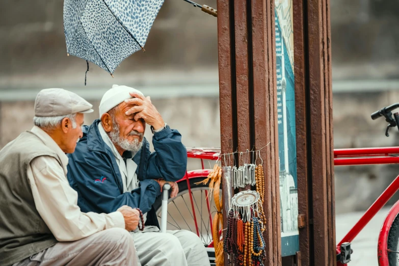 two men sit on the back of a red vehicle under an umbrella