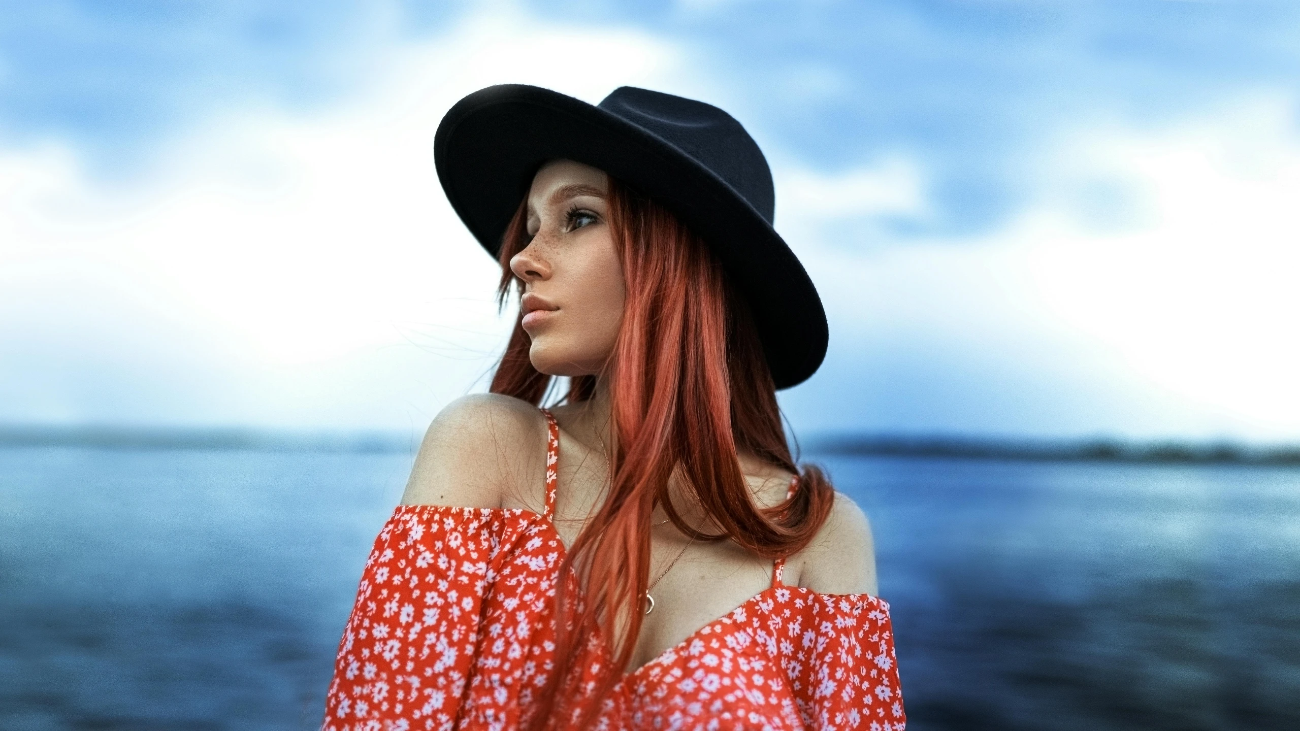 a woman with red hair in a hat and dress standing on a body of water