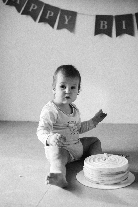 a black and white po of a baby sitting on the floor near a cake