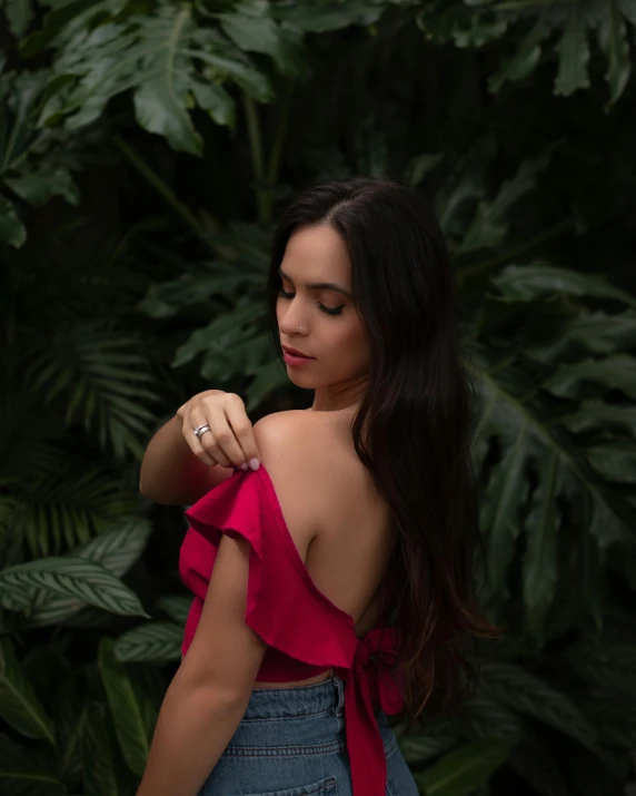 a woman in blue jeans and red top standing near green plants