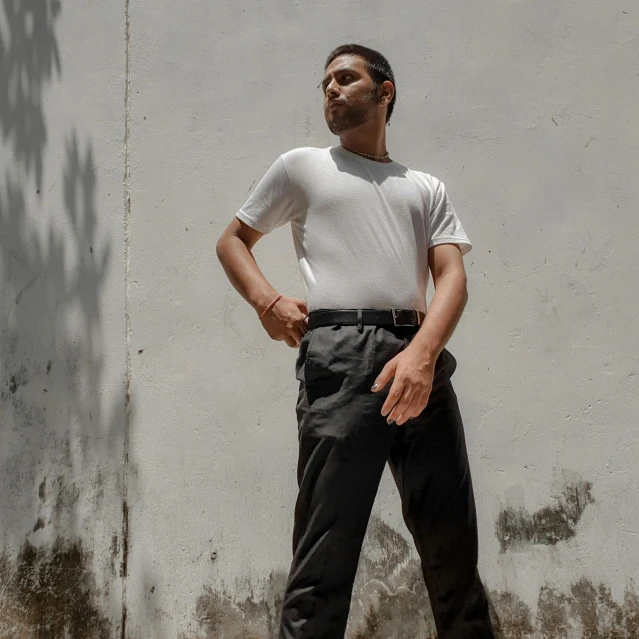 a man leaning against a white wall wearing dark slacks