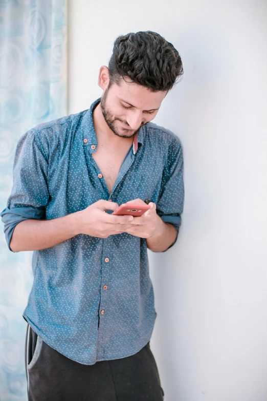 man wearing blue shirt standing next to wall and looking at his cell phone