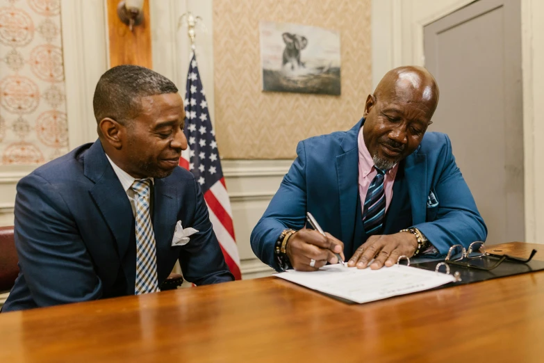 two men in suits are sitting at a wooden table signing a paper