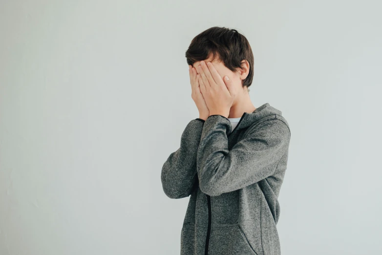 a man covers his face with one hand while standing against a white wall