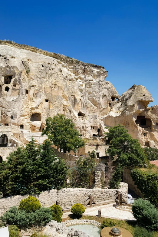 a rock cliff with numerous ruins and trees near a small pool