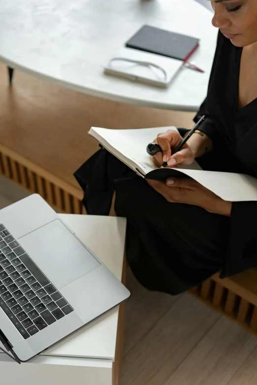 a woman writing in a notebook with a laptop next to her