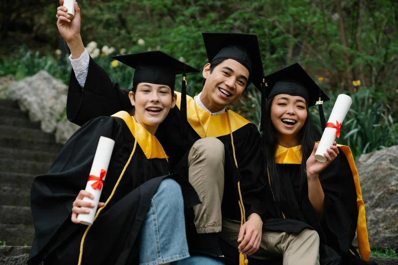 three young men and women sit in front of steps while holding graduation caps