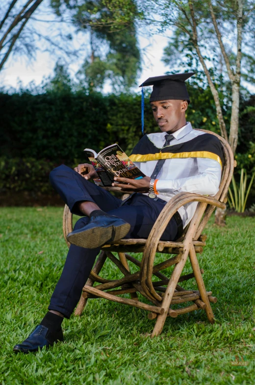 a man in a cap and gown reads a book outside on a lawn chair