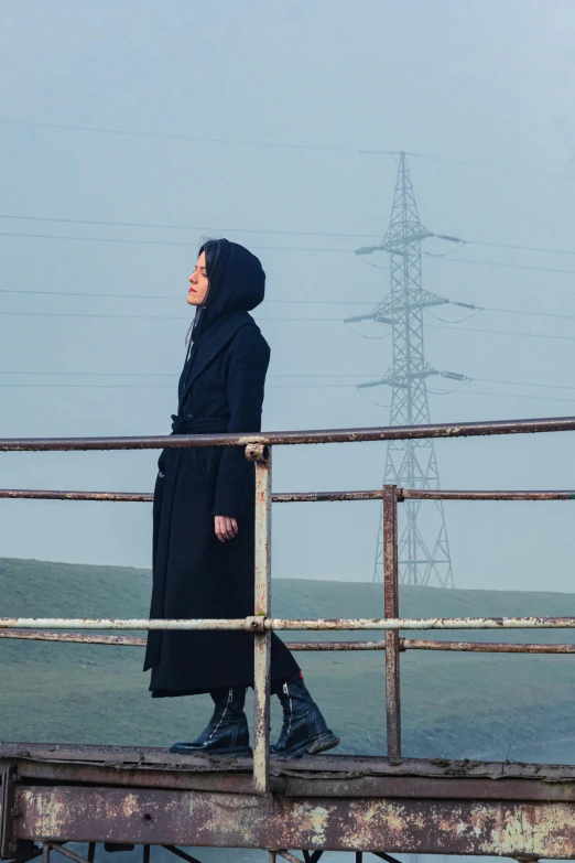 a woman in black clothes and a hood on a bridge overlooking a distant area