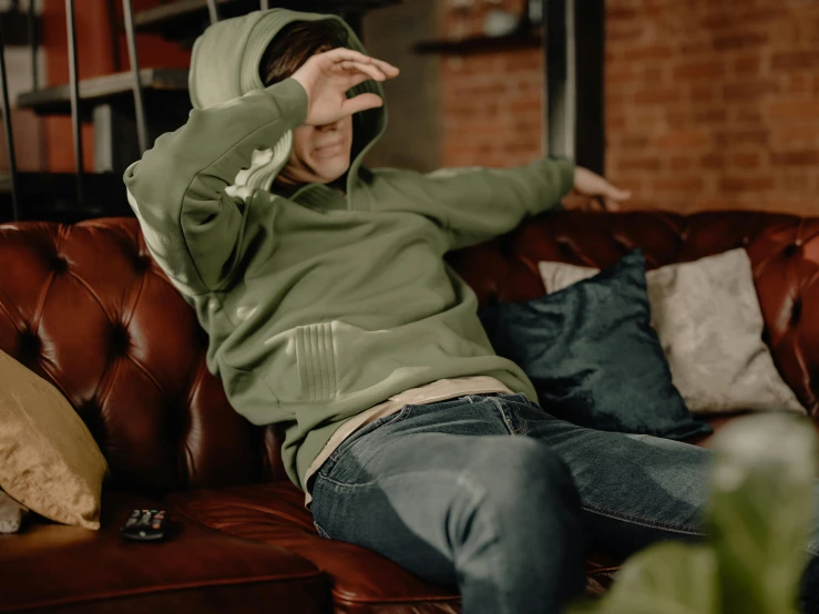 a young man sits on the couch while covering his face with a sweatshirt