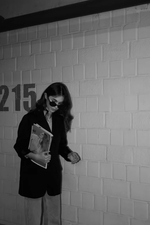 a woman in glasses reading a book standing in front of a brick wall