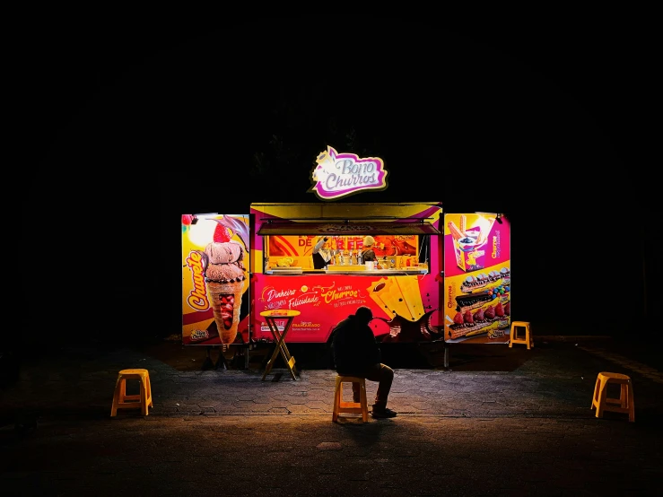 a dark, empty, dark night with a woman sitting at a colorful food cart at night