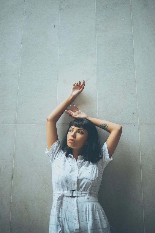 a woman poses in front of a wall with one hand above her head