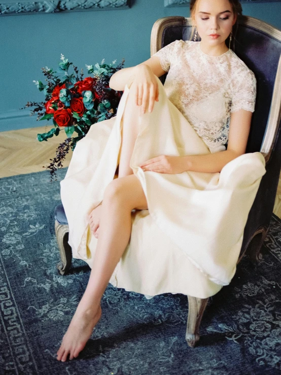 a woman wearing a white dress sitting in an antique chair with a bouquet