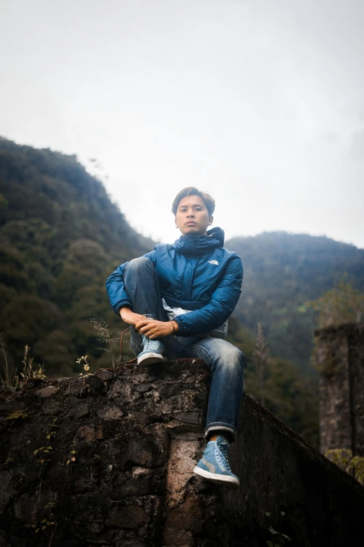 a man in blue jacket sitting on wall by trees