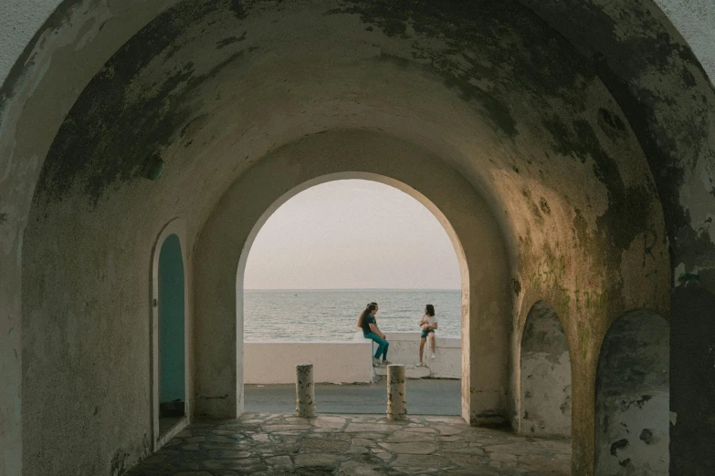 two people standing under a stone arch near the water