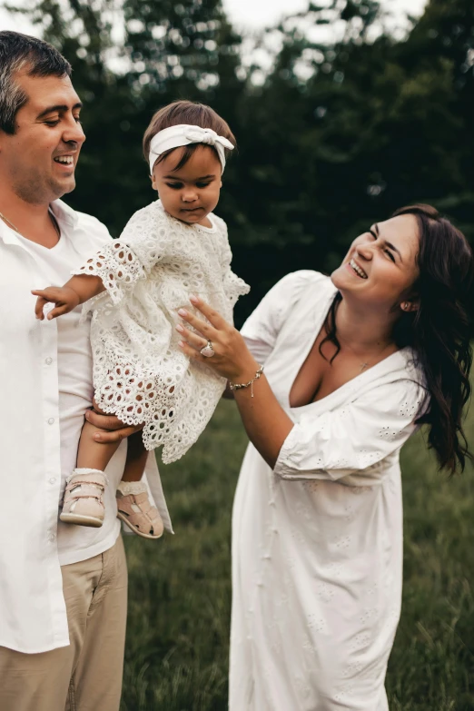 a woman in a white dress holding a baby girl and smiling while the man holds him up