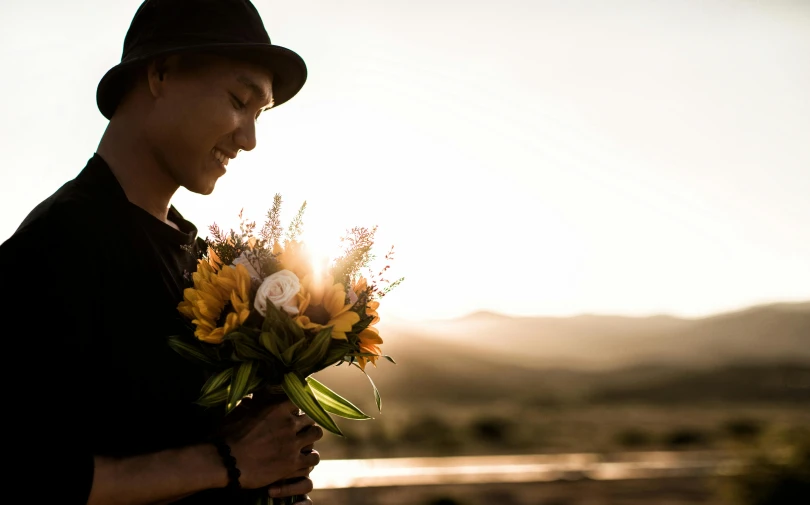 man wearing hat and holding flowers and sun shining behind him