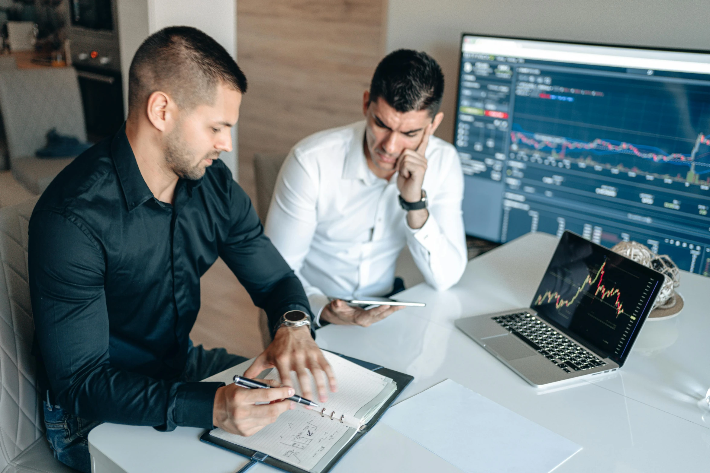 two men sitting at a table, one of them looking at the computer screen