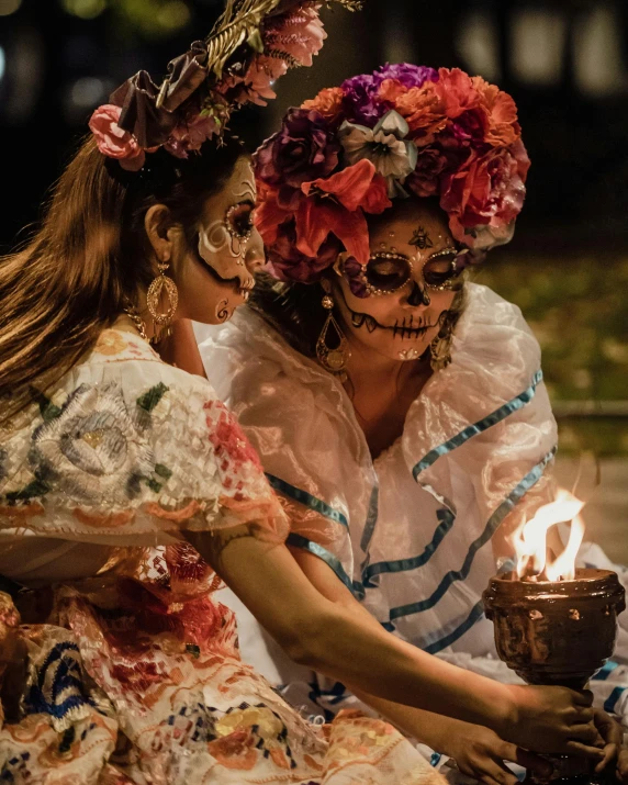 two women wearing sugar skulls and colorful costumes, holding up a lit candle