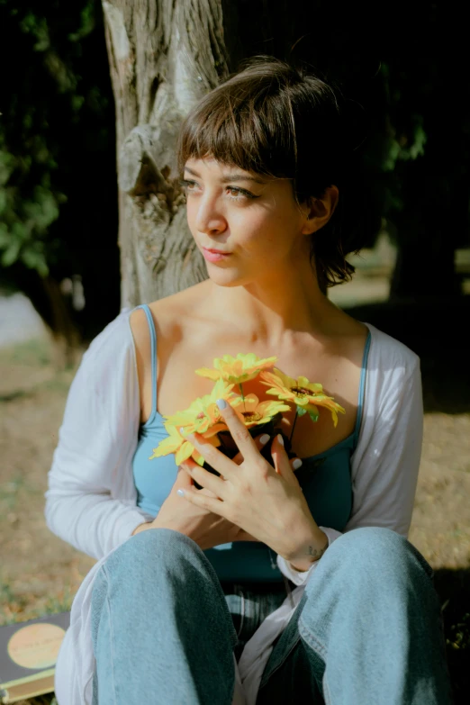 a young woman holding flowers while sitting in front of a tree