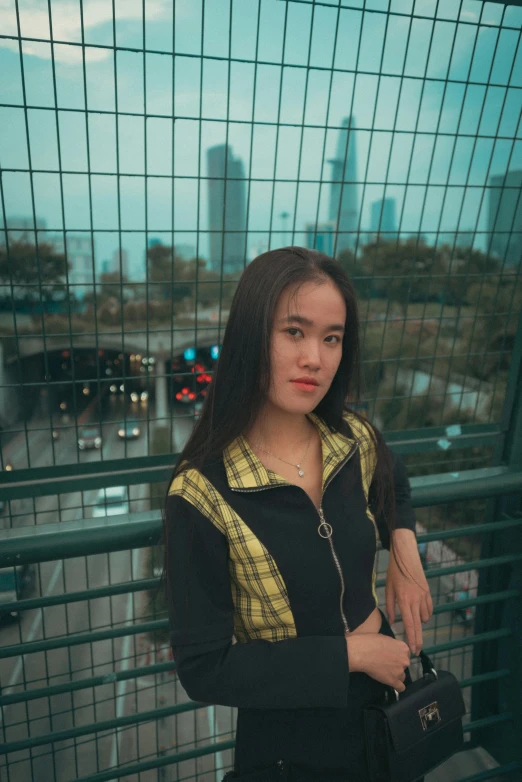 a young asian girl holding a black and gold purse standing on a fence overlooking a city