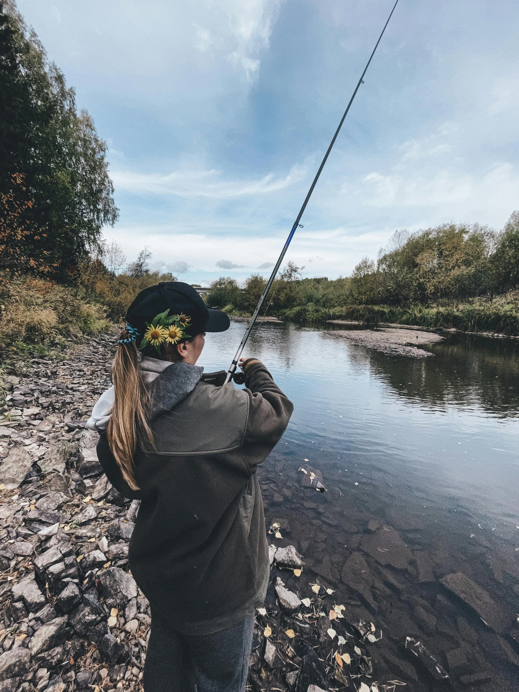 woman fishing on a rocky river bank while wearing a black hat