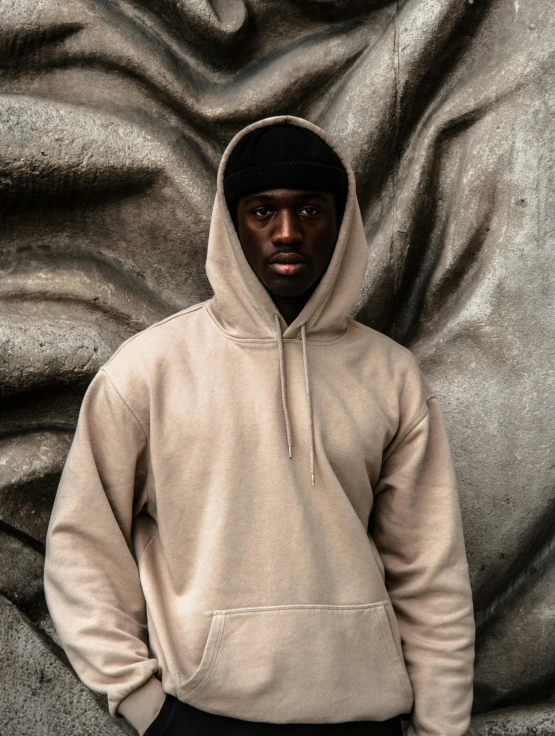 the black man wearing a beige hoodie stands against a silver wall
