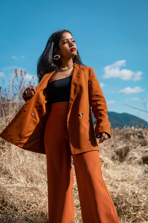 a woman with long hair stands in a field and wears orange pants