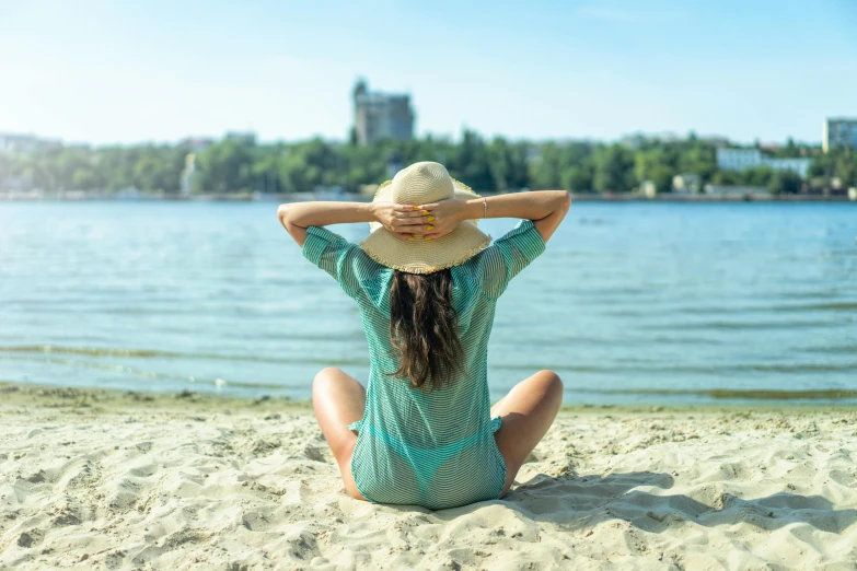 a young woman sitting in the sand at the beach, on top of her hat