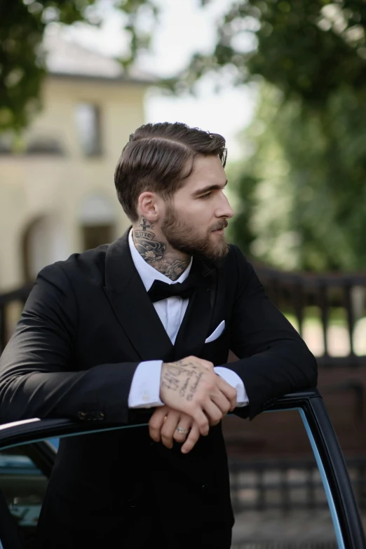 a man with tattoos in a suit leaning on the car door
