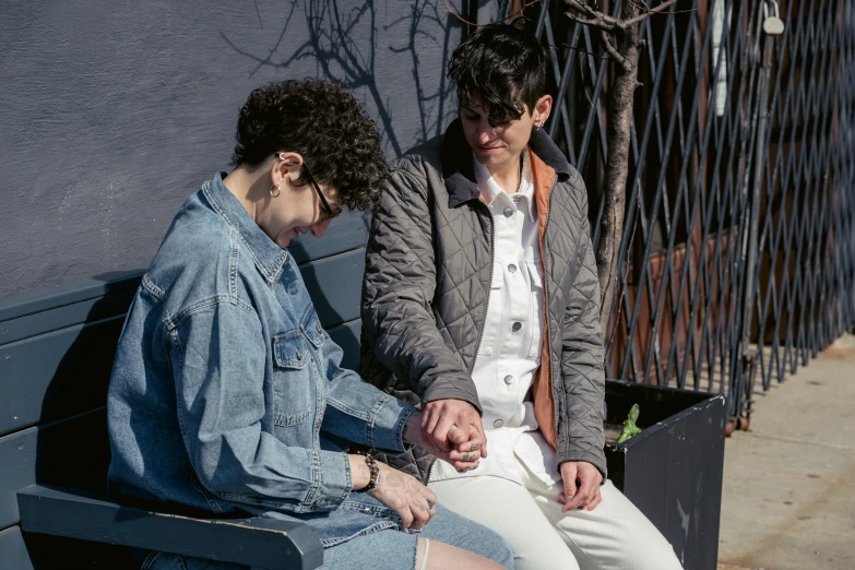 two people sitting on a bench one looks down and one has her foot on the chair