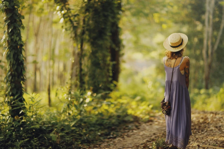 a woman in a long blue dress and straw hat standing in the forest