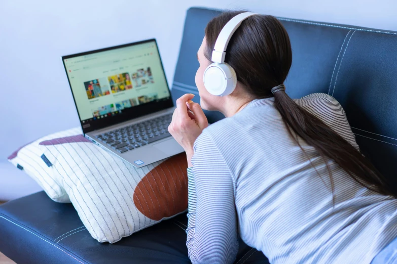 woman on blue leather couch using laptop and wearing headphones