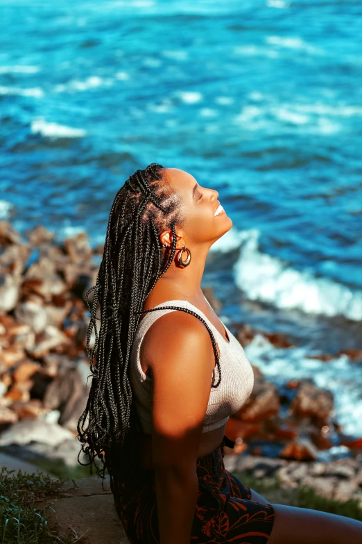 a woman with a black hair sitting on a rocky shore near the water