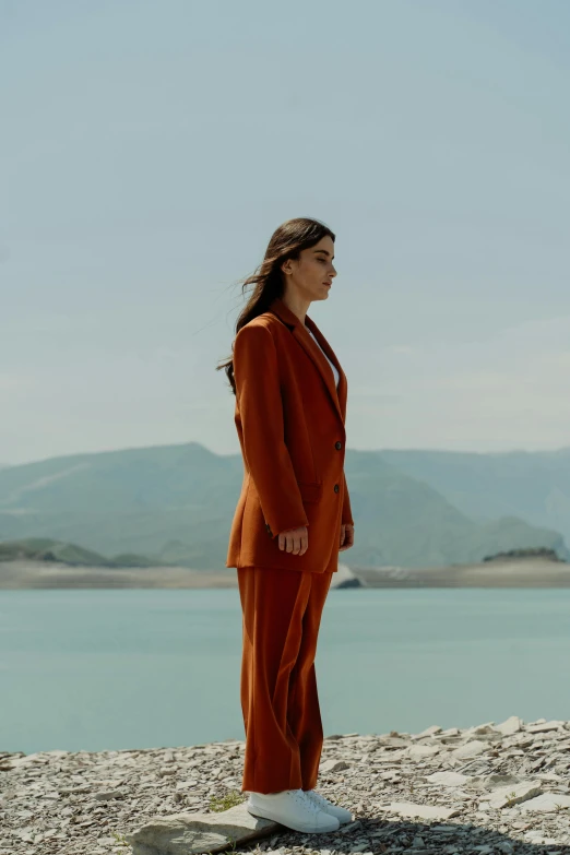 a woman in an orange suit and sneakers, staring out at the water