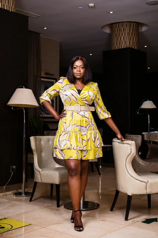 a woman in yellow dress standing beside chair with lamp