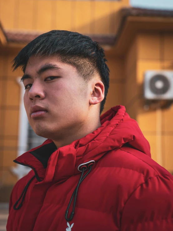 a young man wearing a red parka stares in front of an orange building