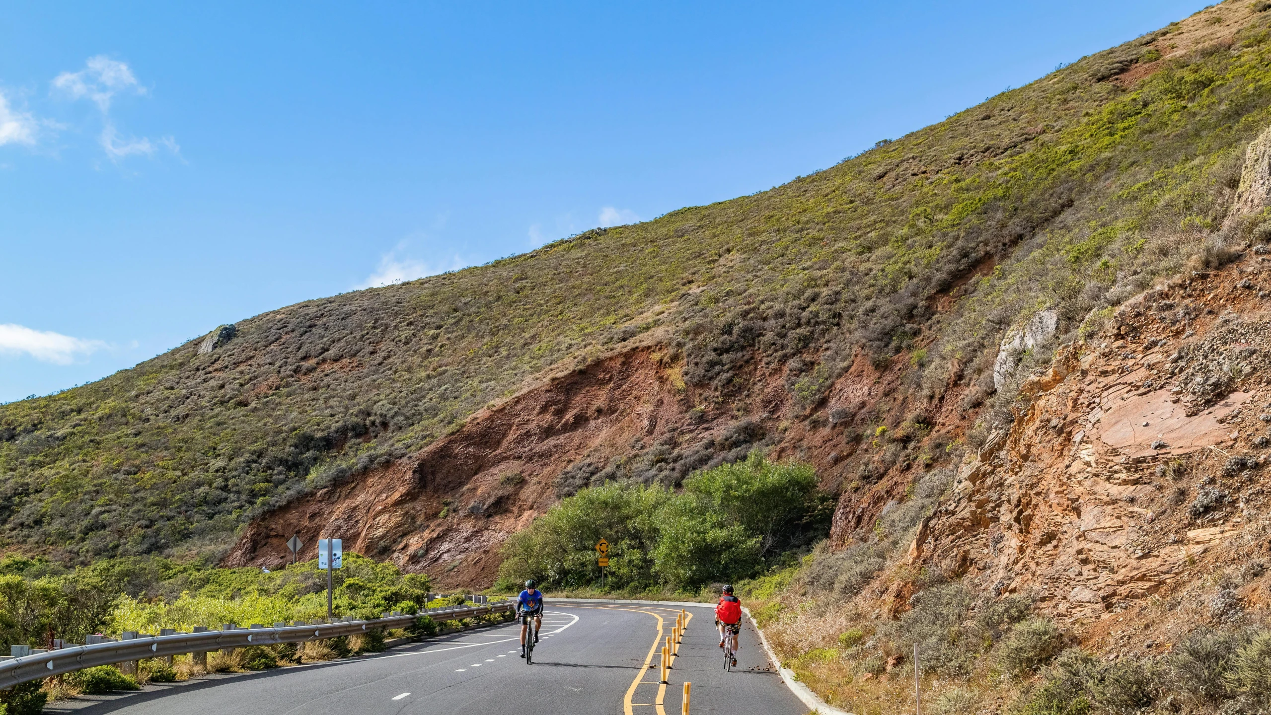 two people walk on a long road by a mountain side