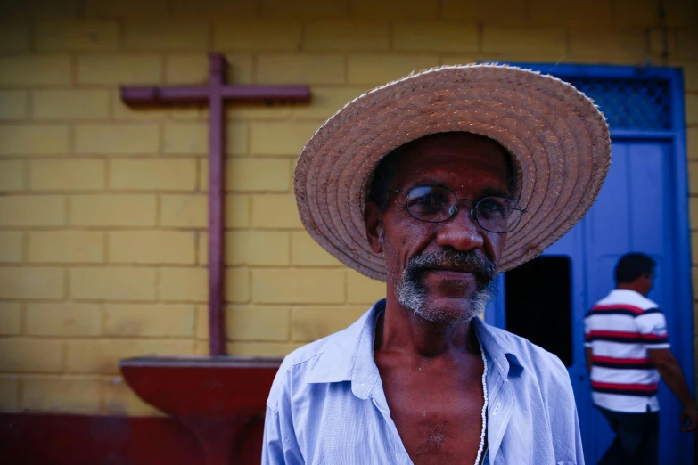 a man with glasses and a straw hat standing by a yellow building