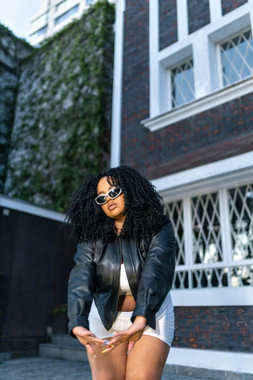 a woman wearing shorts, a white top, and a black leather jacket stands outside of a building