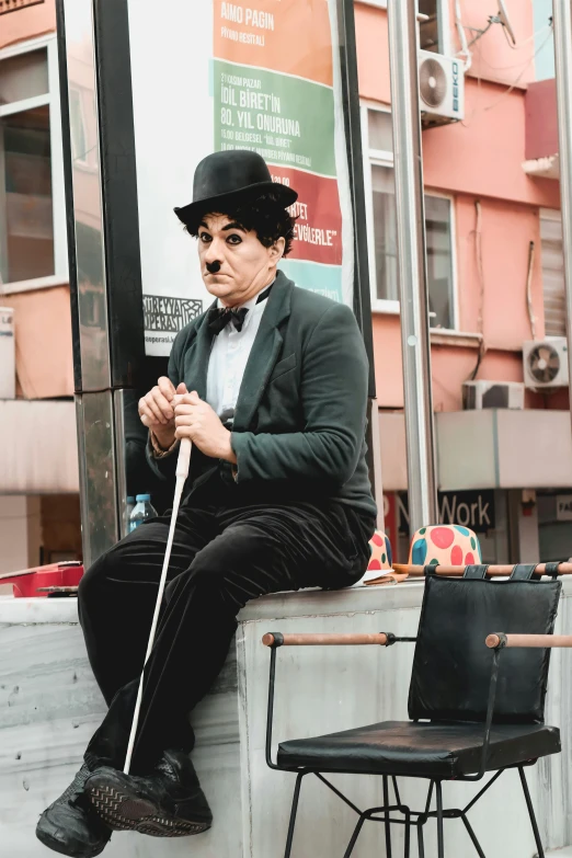 a man with a black suit and fedora sits on a curb near two black chairs and a sign