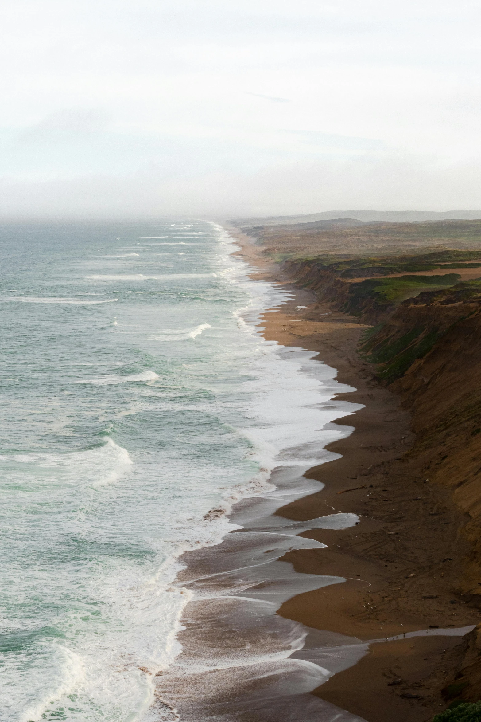 a long sandy beach with a line of cliff and some waves coming towards the shore