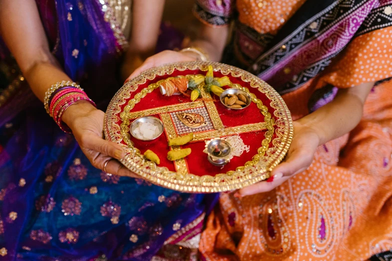 two indian women with brightly colored clothing hold an intricately decorated bowl