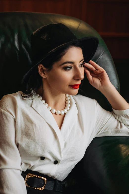 a woman in a black hat, white shirt and pearls sits on a leather chair